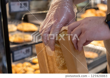 Hand wearing clear plastic glove hygienically packaging freshly baked bread item into brown paper bag at store bakery for convenient customer grab-and-go purchase 125928863