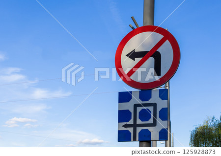 Two traffic signs are mounted on a pole against blue sky, including a No Left Turn symbol and complex diagrammatic sign for road regulation driver guidance Two traffic signs are mounted on a pole against blue sky, including a No Left Turn symbol and complex diagrammatic sign for road regulation driver guidance 125928873