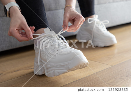 Person's hands tying white laces on chunky sole sneakers indoors on a wooden floor, illustrating preparation for activity and comfortable casual wear 125928885