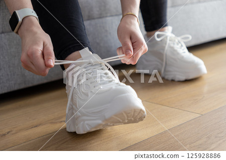 Person's hands adjusting white laces on chunky sole sneakers indoors on a wooden floor, demonstrating fitting for comfortable daily activity Person's hands adjusting white laces on chunky sole sneakers indoors on a wooden floor, demonstrating fitting for comfortable daily activity 125928886