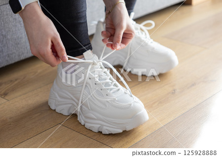 Close up of person's hands tying white laces on chunky sole sneakers indoors, illustrating preparation and fitting for comfortable daily casual wear Close up of person's hands tying white laces on chunky sole sneakers indoors, illustrating preparation and fitting for comfortable daily casual wear 125928888