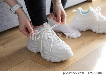 Person's hands fastening white laces on chunky training sneakers indoors, indicating preparation for comfortable and supportive exercise Person's hands fastening white laces on chunky training sneakers indoors, indicating preparation for comfortable and supportive exercise 125928889