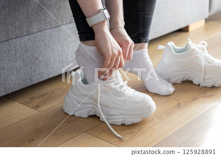 Close up of hands putting on white chunky athletic sneakers indoors, showing preparation for comfortable fitness activity Close up of hands putting on white chunky athletic sneakers indoors, showing preparation for comfortable fitness activity 125928891
