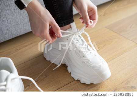 Close up of person's hands tying white laces on chunky training sneakers indoors, preparing for comfortable sport activity Close up of person's hands tying white laces on chunky training sneakers indoors, preparing for comfortable sport activity 125928895