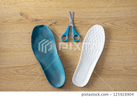 Top view of blue and white insoles with scissors on wooden floor, representing choice and customization for comfortable athletic footwear support Top view of blue and white insoles with scissors on wooden floor, representing choice and customization for comfortable athletic footwear support 125928904
