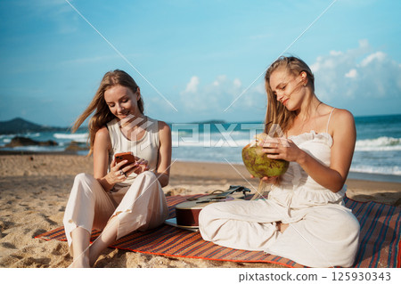 Young Women Smiling on the Beach with Drinks and Smartphones Young Women Smiling on the Beach with Drinks and Smartphones 125930343