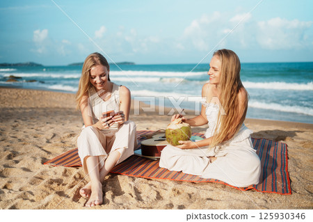 Two Women Relaxing on the Beach with Refreshing Coconut Two Women Relaxing on the Beach with Refreshing Coconut 125930346