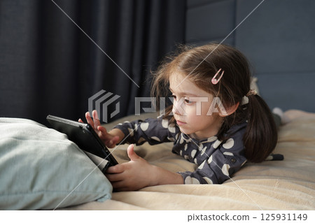 Focused little girl lies on bed and interacts with tablet screen, her face serious and intent 125931149