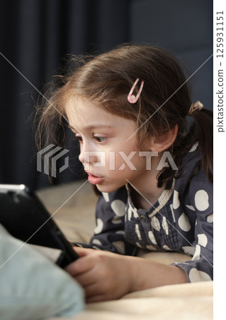 Little girl intently watching tablet screen with surprised expression while lying on bed 125931151