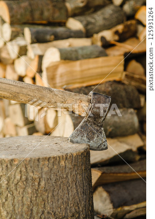 An axe is stuck in a sturdy stump. Behind it, there are many chopped and stacked logs. An axe is stuck in a sturdy stump. Behind it, there are many chopped and stacked logs. 125931264