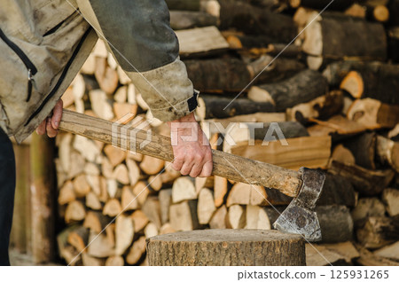 A firm hand holds an axe embedded in a stump. Stacks of chopped wood rest in the background. A firm hand holds an axe embedded in a stump. Stacks of chopped wood rest in the background. 125931265