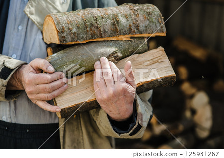 Rough, veined hands carry freshly chopped logs. Behind them, firewood is piled high. 125931267