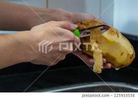 Potapo peeling. Hands peeling raw potato with vegetable peeler in the kitchen.	 125933538