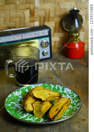 Vintage still life with Pisang Rebus or steamed bananas on a green-white plate, coffee in a glass mug, antique radio, and red-gold oil lamp on wooden surface. 125933869