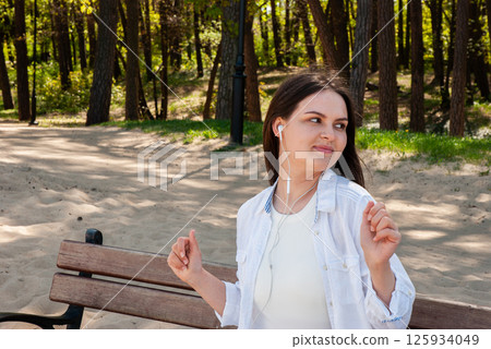 Young caucasian female enjoying music with earphones on a sunny day in a green park, seated on a wooden bench amidst tall trees. Digital nomad lifestyle. Remote work Young caucasian female enjoying music with earphones on a sunny day in a green park, seated on a wooden bench amidst tall trees. Digital nomad lifestyle. Remote work 125934049
