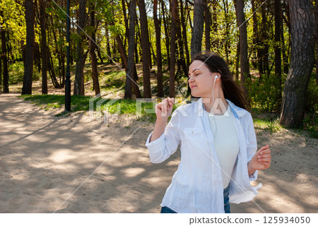 Young caucasian female in white shirt joyfully dancing outdoors with earphones among trees in sunlit park path. Digital nomad lifestyle. Remote work Young caucasian female in white shirt joyfully dancing outdoors with earphones among trees in sunlit park path. Digital nomad lifestyle. Remote work 125934050