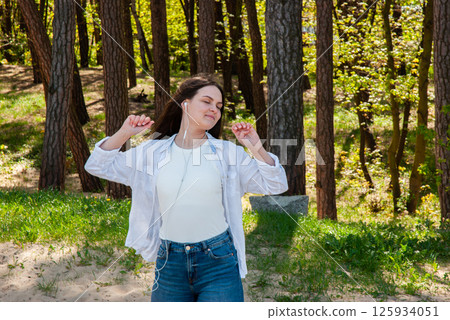 Young caucasian woman enjoying music outdoors in a serene forest setting, wearing casual attire with a relaxed expression and earphones. Digital nomad lifestyle. Remote work Young caucasian woman enjoying music outdoors in a serene forest setting, wearing casual attire with a relaxed expression and earphones. Digital nomad lifestyle. Remote work 125934051