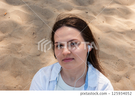 Young caucasian female with earphones at beachside, engaged in relaxing moment, wearing casual attire, surrounded by sandy shores. Digital nomad lifestyle. Remote work 125934052