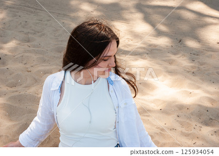 Young caucasian woman with long brown hair enjoying a sunny day at the sandy beach, wearing casual white attire and listening to music. Digital nomad lifestyle. Remote work 125934054