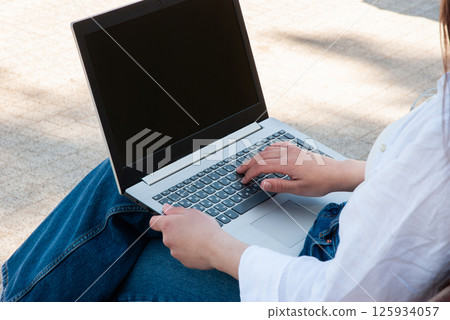 Young caucasian female sitting outdoors focused on her laptop wearing a white shirt and denim jeans under natural sunlight on a sunny day. Digital nomad lifestyle. Remote work Young caucasian female sitting outdoors focused on her laptop wearing a white shirt and denim jeans under natural sunlight on a sunny day. Digital nomad lifestyle. Remote work 125934057
