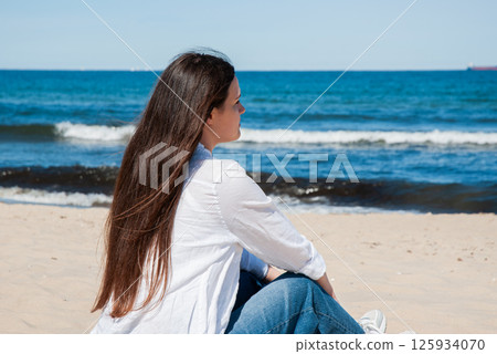 Young caucasian female with long brown hair in casual attire sitting on a sandy beach, gazing at the calm ocean waters under a clear blue sky. Digital nomad lifestyle. Remote work 125934070