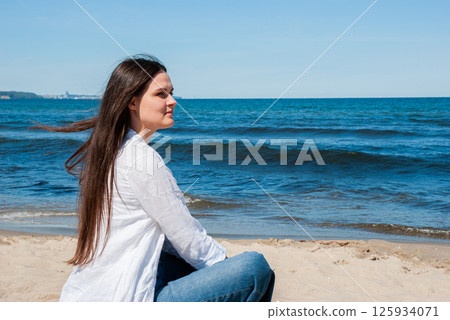 Young caucasian female sitting on sandy beach in casual clothing with long brown hair, blue jeans, and white shirt by clear blue ocean waves. Digital nomad lifestyle. Remote work 125934071