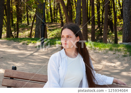 Young caucasian female relaxing on park bench in forested area during a bright day, wearing white shirt and looking thoughtful and serene. Digital nomad lifestyle. Remote work Young caucasian female relaxing on park bench in forested area during a bright day, wearing white shirt and looking thoughtful and serene. Digital nomad lifestyle. Remote work 125934084