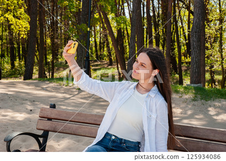 Young caucasian female taking a selfie on a sunny day in a park, sitting on a wooden bench surrounded by vibrant green trees and sandy ground. Digital nomad lifestyle. Remote work Young caucasian female taking a selfie on a sunny day in a park, sitting on a wooden bench surrounded by vibrant green trees and sandy ground. Digital nomad lifestyle. Remote work 125934086