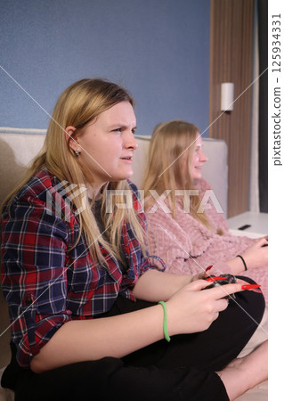 Two teen girls intensely focused on a video game, one showing frustration and concentration while the other smiles slightly, both holding controllers in a cozy indoor setting. 125934331