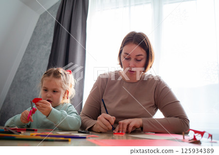 Low angle view of adult Caucasian mother and her little daughter cut out paper hearts sitting at a desk. Family is preparing for the celebration of Valentine's Day Low angle view of adult Caucasian mother and her little daughter cut out paper hearts sitting at a desk. Family is preparing for the celebration of Valentine's Day 125934389