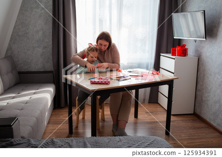 Wide shot of young woman teacher helps a little girl to cut out paper hearts for the Valentine's Day holiday. Concept of children education 125934391