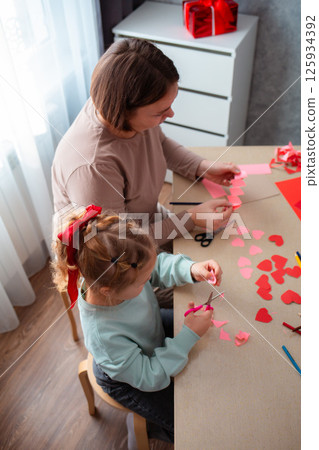 Top view of little cute girl and her mother cut out paper hearts sitting at a desk. Family is preparing for the celebration of Valentine's Day, Vertical 125934392