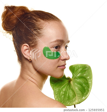 Side view image of beautiful young redhaired woman with natural skin and freckles, in eye patch, with green leaf over white background. 125935951