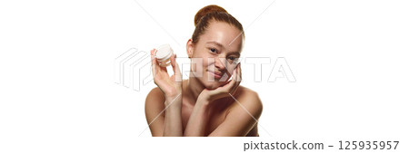 Portrait of beautiful young woman with spotless freckled skin holding jar of cream, touching cheek over white background. 125935957