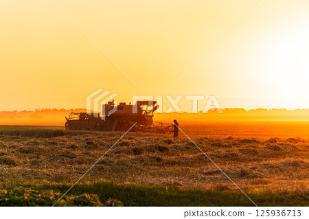 A farmer operates a harvesting machine in a golden field at sunset, capturing the essence of rural life and hard work 125936713
