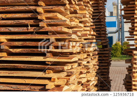 Stacks of neatly arranged wooden planks are seen in a lumber yard, illuminated by bright daylight against a clear sky 125936718