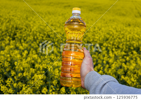 A hand holds a bottle of canola oil in a vast field of blooming rapeseed flowers, illustrating the connection between farming and the environment 125936775