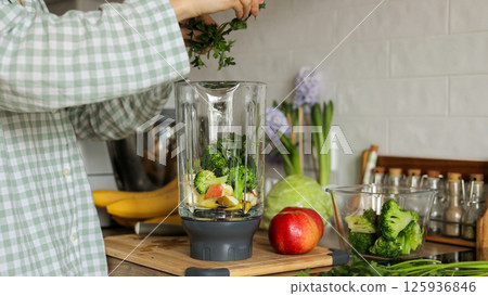 Unknown woman makes healthy detox breakfast smoothie at home. Female hands throws parsley in blender bowl, prepares fresh green vegetarian fruit-greens-vegetables drink to cleanse, provide energy body Unknown woman makes healthy detox breakfast smoothie at home. Female hands throws parsley in blender bowl, prepares fresh green vegetarian fruit-greens-vegetables drink to cleanse, provide energy body 125936846