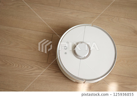 Top view of white wireless autonomous robotic vacuum cleaner on wooden floor. Smart cleaning technology. Self-propelled cleaning robot. Floor cleaning system. Household remote control appliances. 125936855