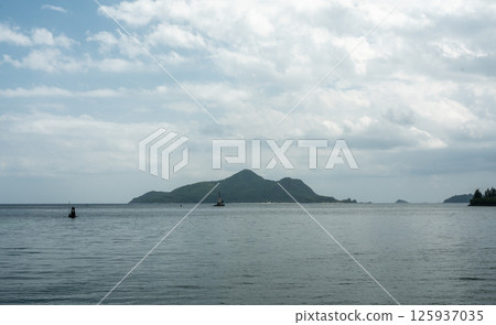 Mahe island in Seychelles seen from a boat, lush green hills and calm sea under cloudy sky 125937035
