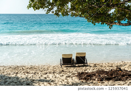 Two lounge chairs on a tropical Seychelles beach facing calm turquoise waves under sunny skies 125937041