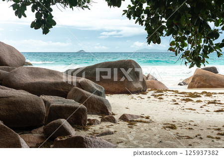 Tropical beach with large boulders, turquoise ocean waves, distant islands and lush vegetation Tropical beach with large boulders, turquoise ocean waves, distant islands and lush vegetation 125937382