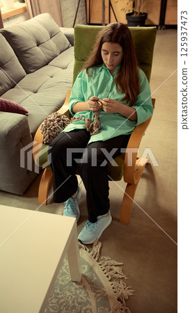 Teenage girl sitting in chair, engaged in knitting with focused attention on the activity highlights her. Cozy living room interior. Teenage girl sitting in chair, engaged in knitting with focused attention on the activity highlights her. Cozy living room interior. 125937473