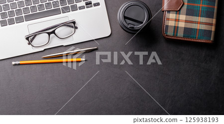 Top view of a dark leather office desk with laptop, supplies, and coffee cup 125938193