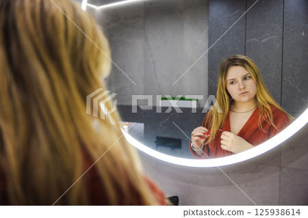 Young woman in a red robe looks thoughtfully at her reflection in a round backlit mirror while holding a toothbrush in a modern bathroom. 125938614