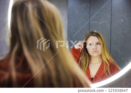 Young woman in a red robe stands in front of a glowing round mirror, thoughtfully examining her reflection while holding a toothbrush in a stylish bathroom. 125938647