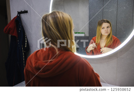 Young woman in a red bathrobe stands at the sink, touching her hair while thoughtfully examining her reflection in a round illuminated mirror. 125938652