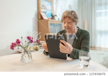 A senior woman uses tablet while sitting at a table in a cozy home environment. Concept of technology, digital literacy, online communication, independent learning, and active aging. 125938670