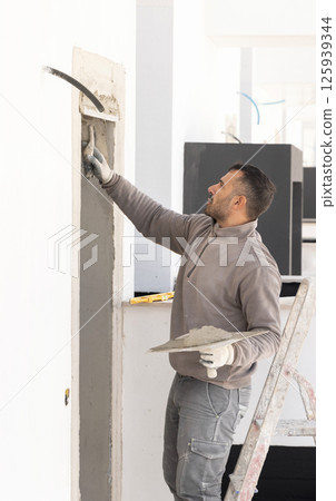 Craftsman in dirty gloves and gray clothes spreading plaster with a metal float over a doorway 125939344
