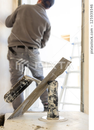 Craftsman in grey clothes working to a doorway during construction, metal trowels in the foreground 125939346